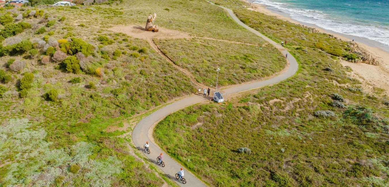 Aerial view of a coastal bike path winding through green and brown vegetation. Four cyclists ride along the path. The ocean is visible on the right, with sandy beach and blue waves. A wooden sculpture and a small group of people are near the path.