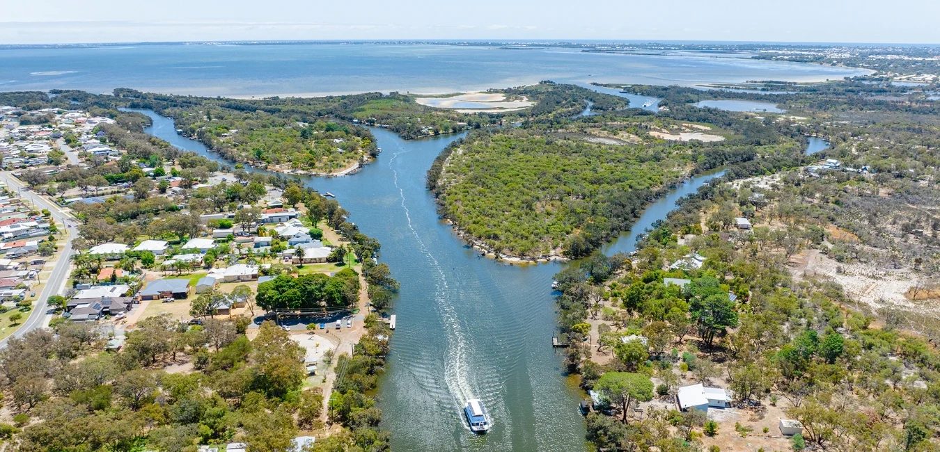 Aerial view of a river winding through lush green forests with a boat on the water. Residential areas are visible on the left, and the river opens into a wide bay in the background under a clear blue sky.