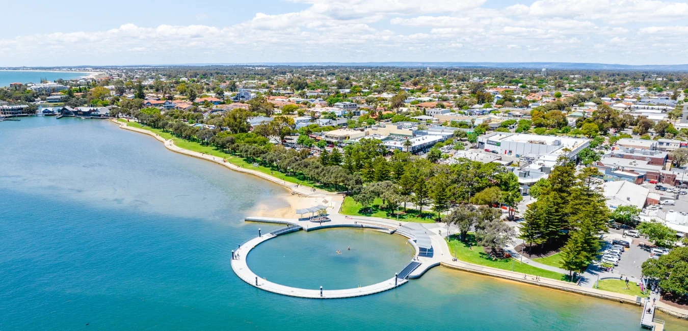 Aerial view of a coastal town with a circular promenade extending into the bay. The shoreline is lined with trees and grass. Residential and commercial buildings are visible in the background under a partly cloudy sky.