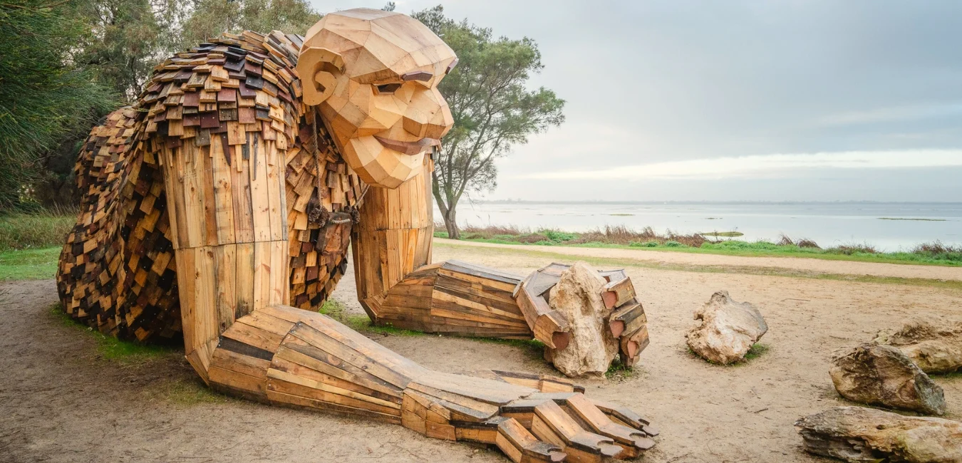 A large wooden sculpture of a troll-like figure crouches beside a path near the estuary The sculpture is crafted from various pieces of wood, and trees are visible in the background under a cloudy sky.