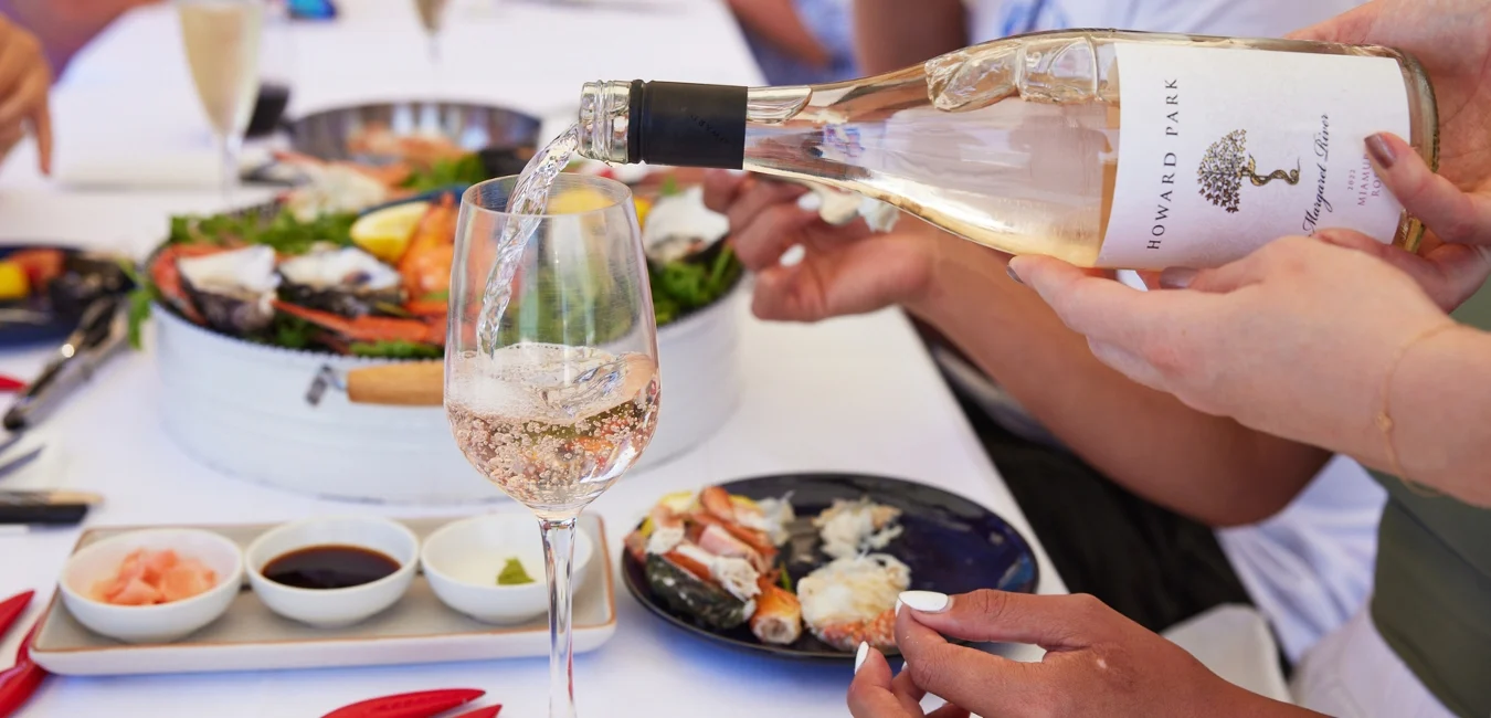 A close-up of a person pouring rosé wine from a Howard Park bottle into a glass. The background features a seafood platter and various dipping sauces on a white tablecloth. Several people are seated around the table.