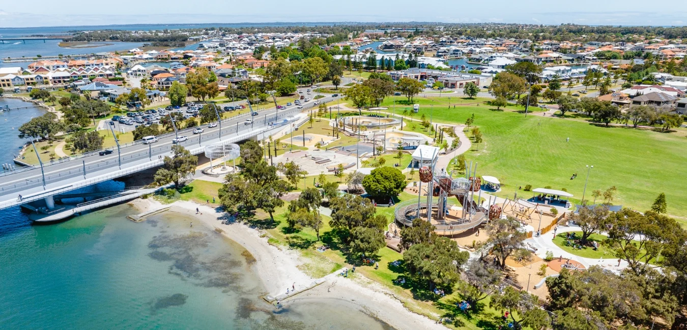 Aerial view of a coastal park with a playground, lawns, and walking paths near a bridge. Residential area and water body visible in the background. People are enjoying the sunny day in the park.