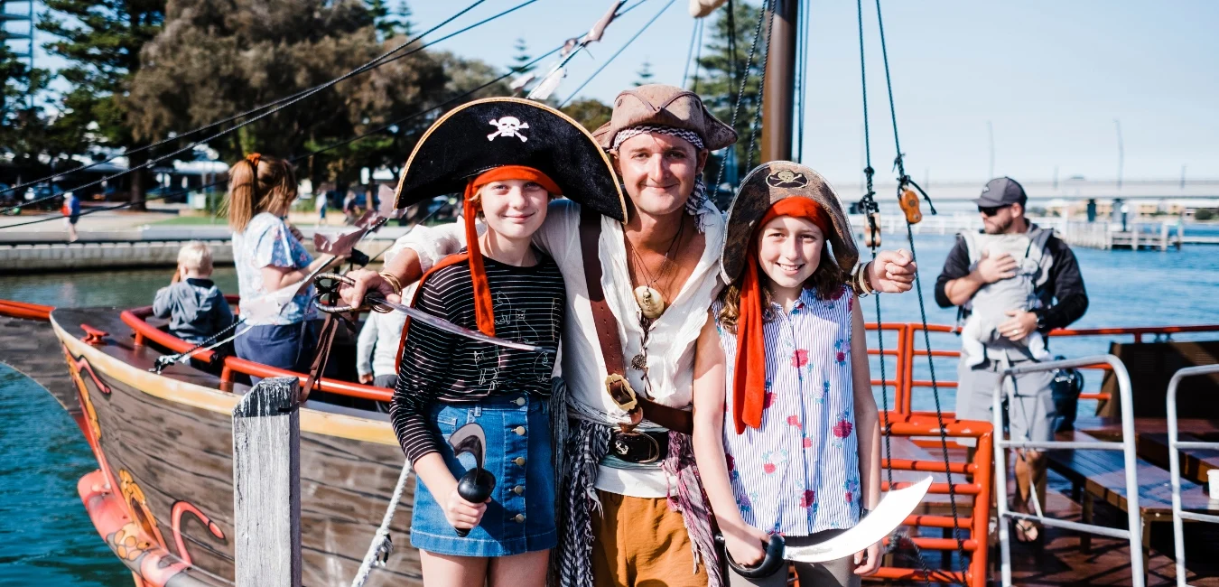 A man and two children dressed as pirates pose in front of a wooden ship by the water. The man wears a brown hat and holds a sword, while the children, in pirate hats, hold toy swords. People and trees are visible in the background.