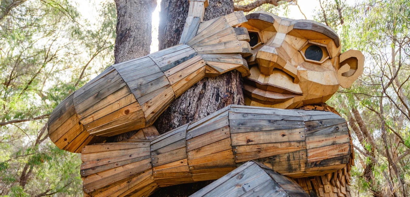 A large wooden sculpture of a troll with exaggerated features, hugging a tree trunk. The sculpture is made from reclaimed wood and set in a forested area with sunlight filtering through the leaves.