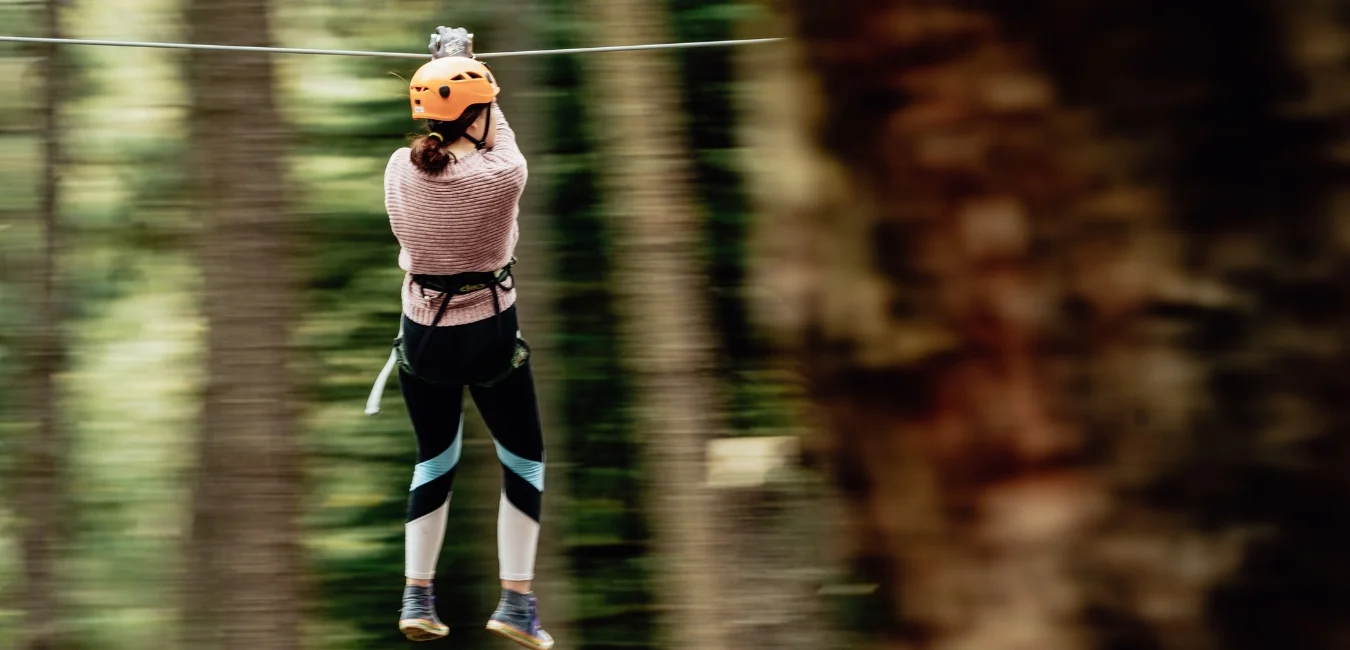 A person wearing a helmet and harness is ziplining through a forest. They are dressed in a pink sweater and black leggings with blue stripes. The background is a blur of green and brown trees.