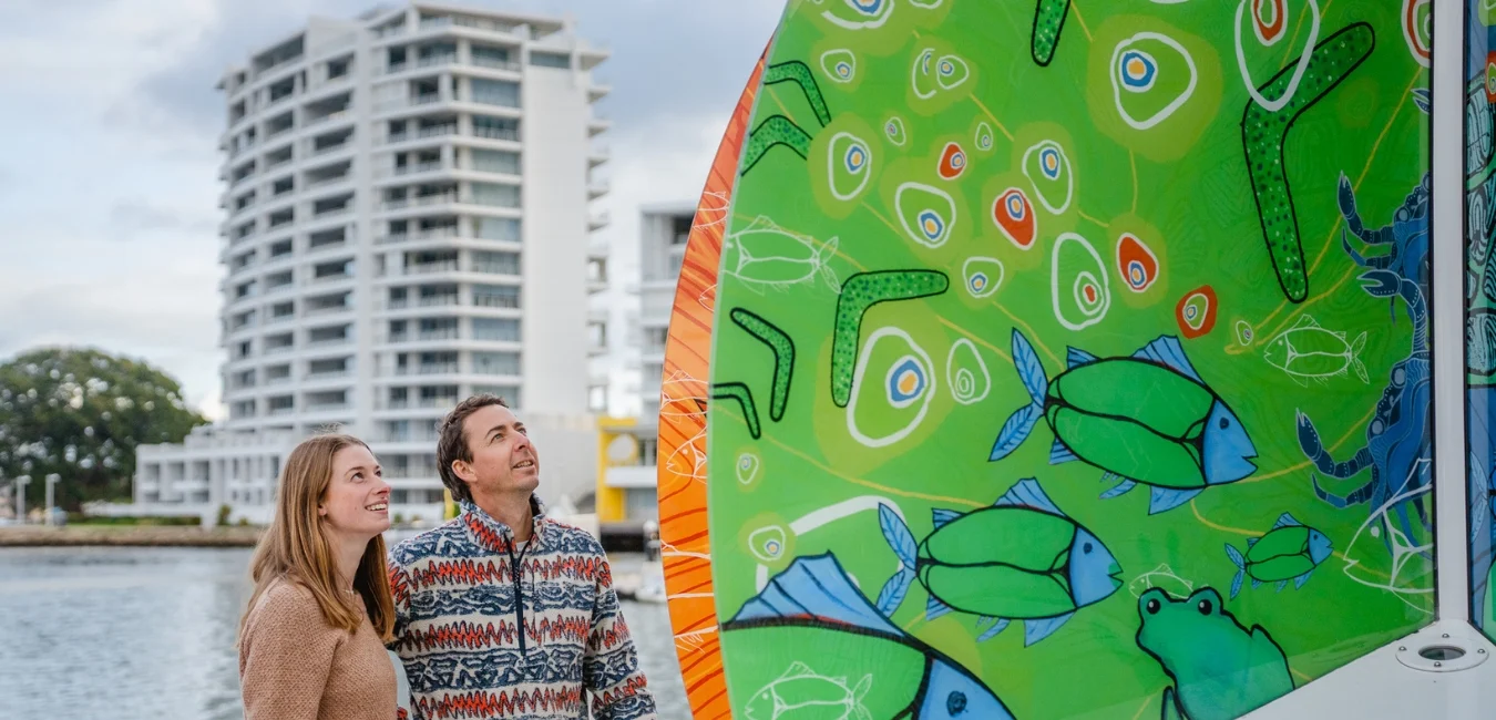 A man and woman stand near a colourful Aboriginal artwork. They are looking up, with residential buildings and water in the background.