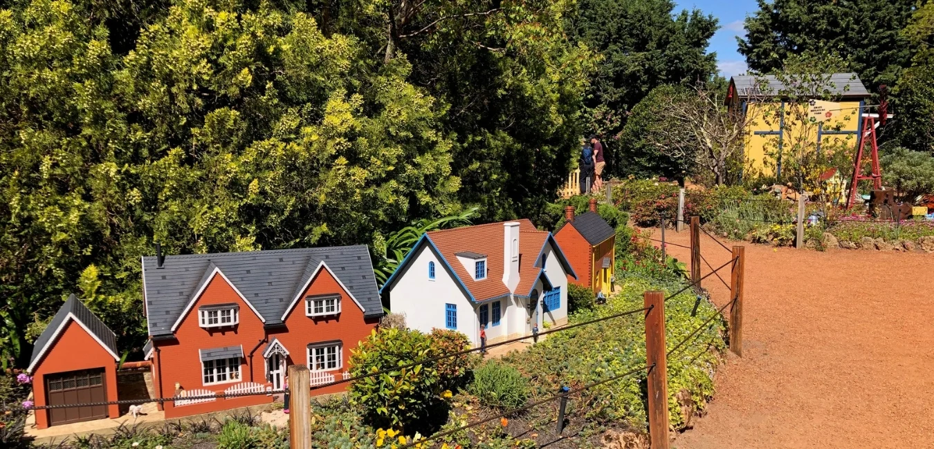 A miniature village with small, detailed model houses surrounded by lush greenery and plants. A dirt path runs alongside, with trees and a person visible in the background. The sky is clear and sunny.