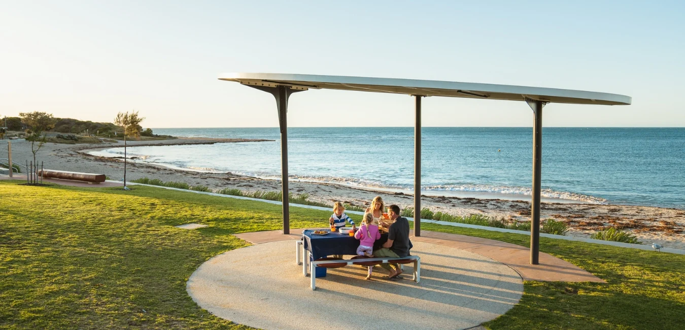 A family of four sits at a picnic table under a modern shelter by a beach. They are enjoying a meal with the ocean and coastline in the background, during a sunny day. The grassy area around them is well-maintained.
