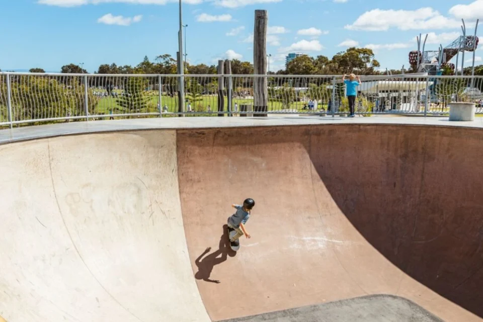 A skateboarder wearing a helmet rides in a large, sunlit skate bowl. A person stands at the edge watching. The backdrop features a park with trees under a blue sky with clouds.