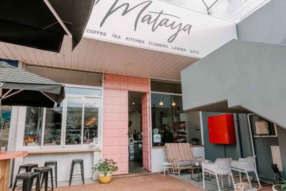 Outdoor view of Mataya café with a welcoming entrance. Black stools line an outdoor counter under an umbrella. White chairs and a striped sofa sit at the entrance. A pink door and sign with Coffee Tea Kitchen Flowers Larder Gifts are visible.