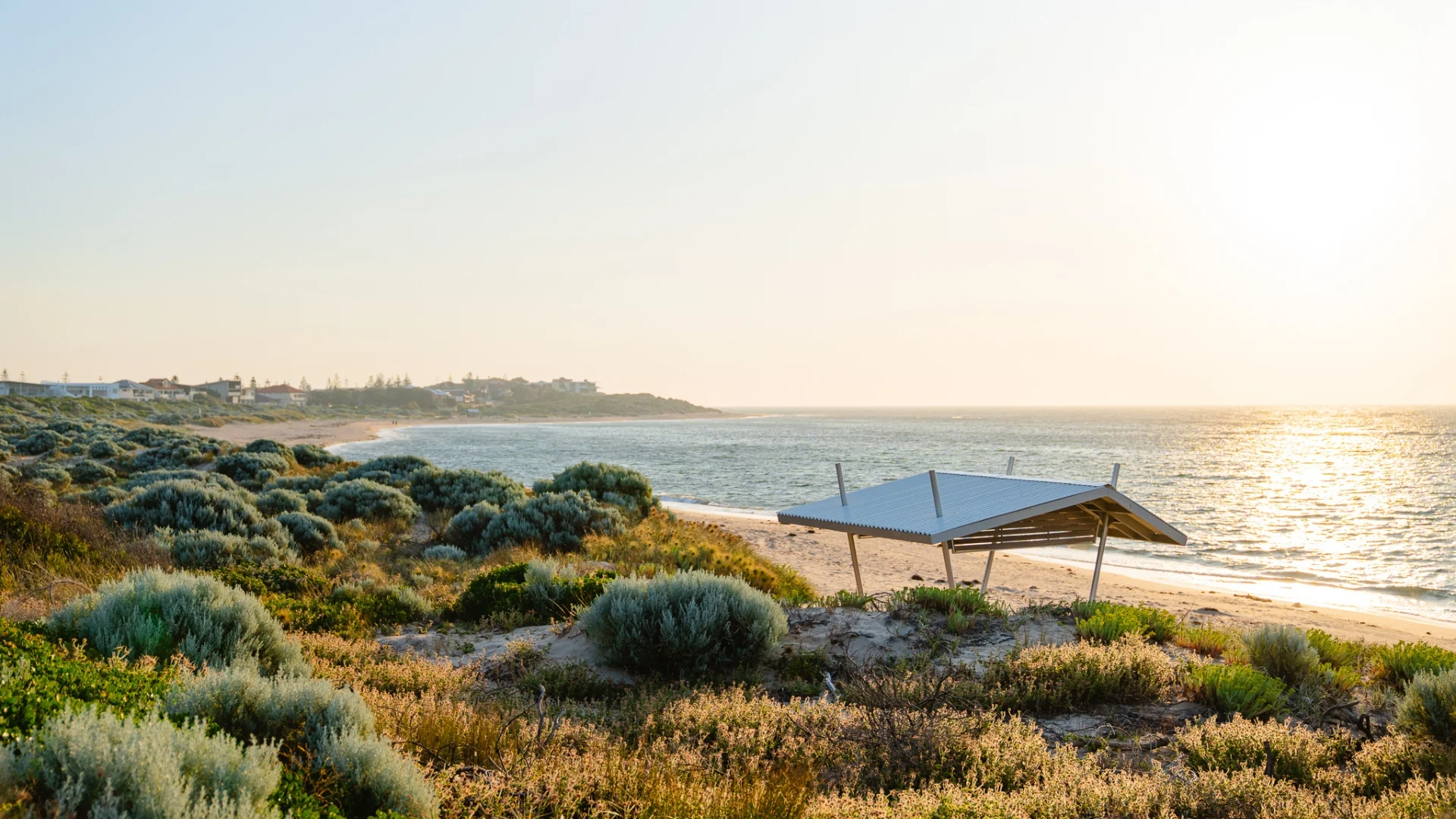 A sandy beach with a small open shelter, surrounded by green shrubs and overlooking calm ocean waves at sunset. The sun casts a golden glow over the landscape and distant buildings are visible on the horizon.
