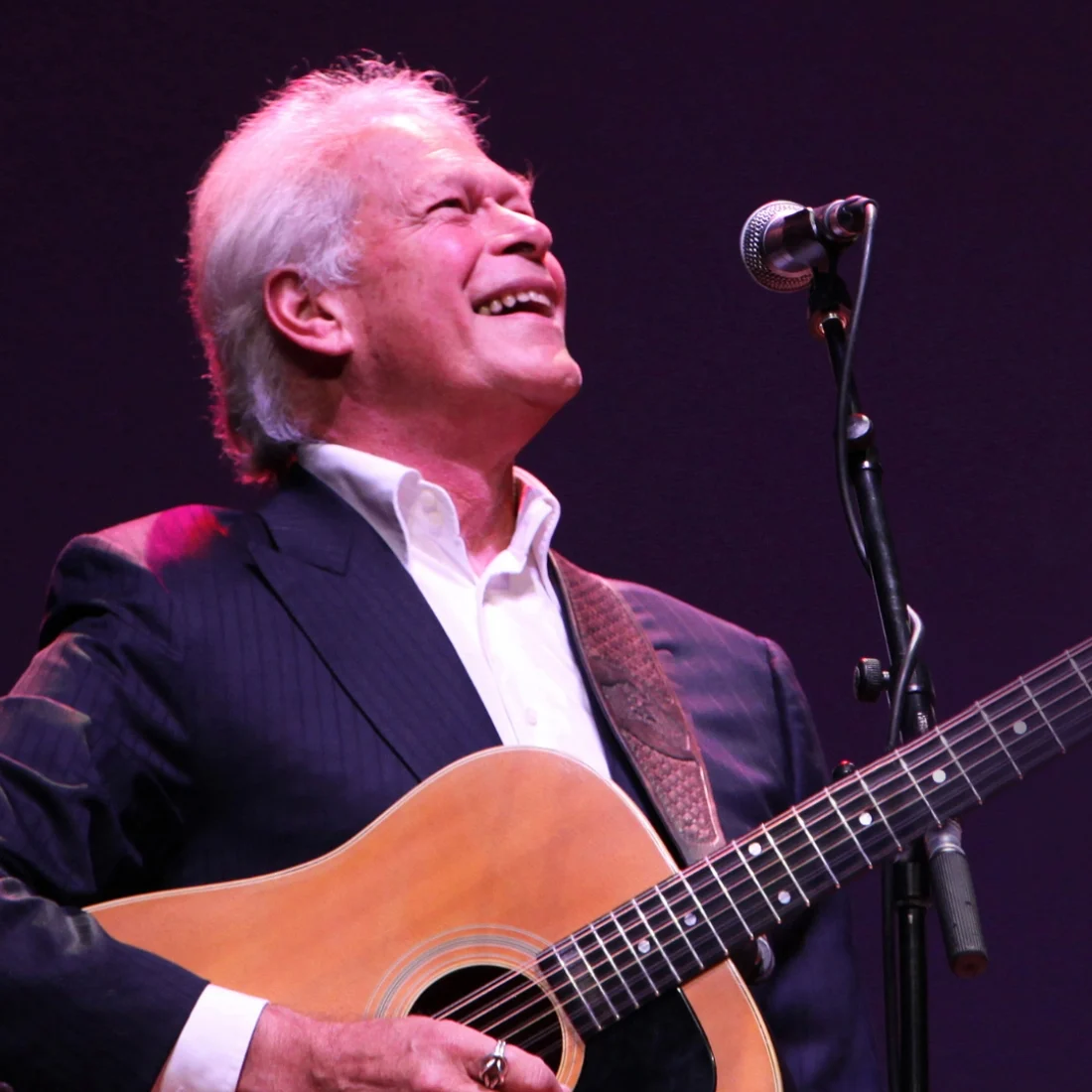 An older man with white hair, wearing a dark suit and white shirt, smiles while playing an acoustic guitar and singing into a microphone on stage.