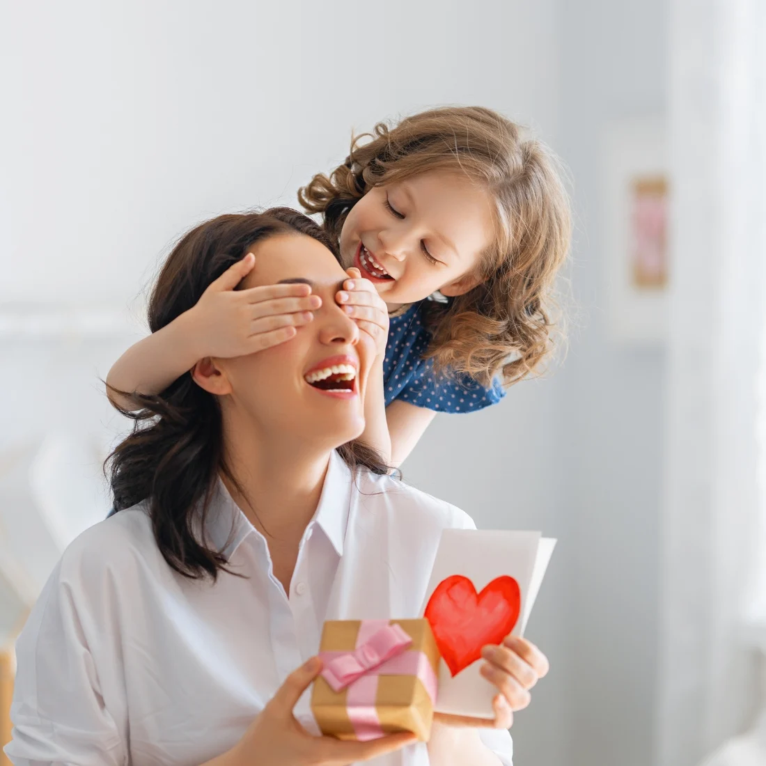 A smiling woman holds a gift and a card with a red heart while a young girl playfully covers her eyes from behind in a bright, cosy room.