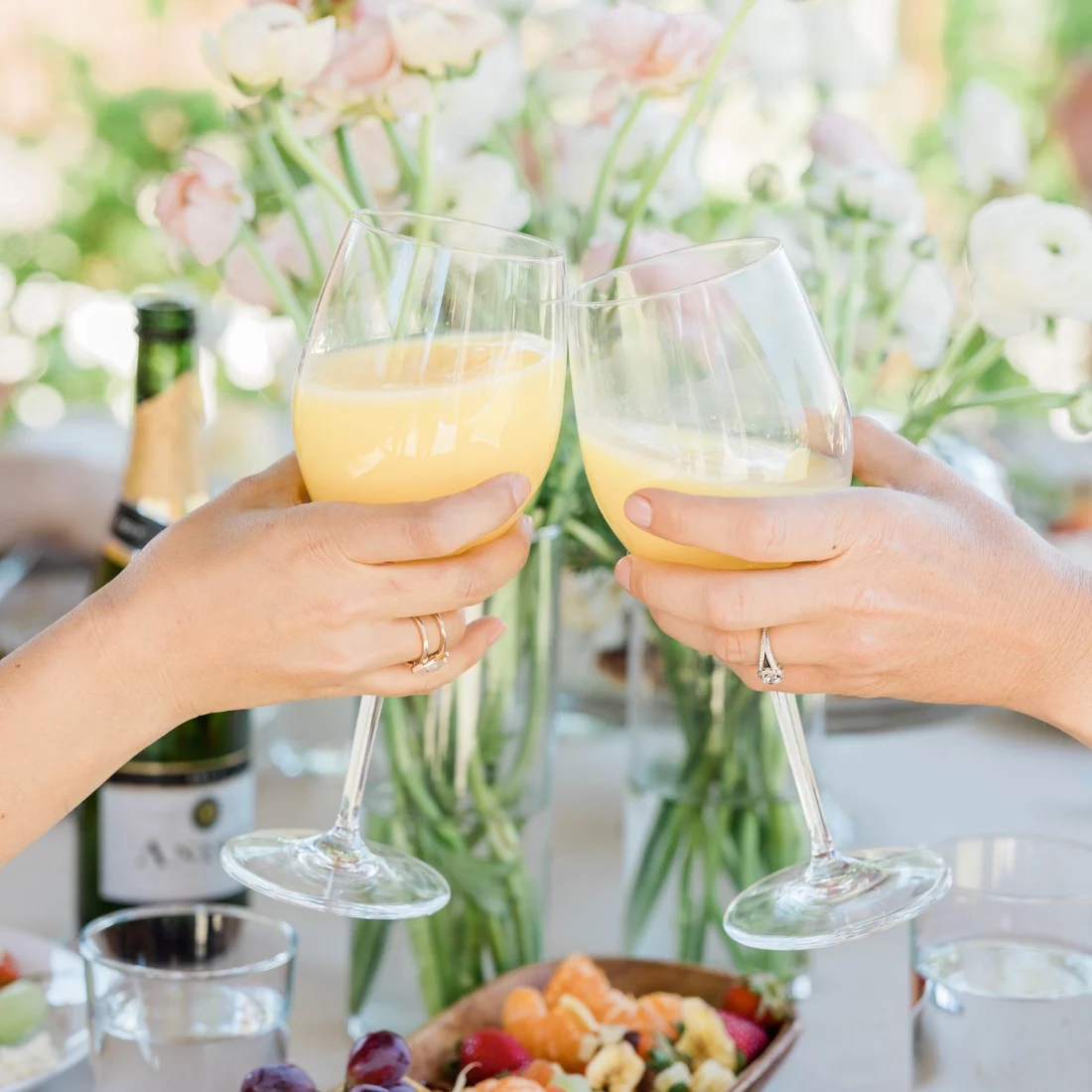 Two people clink glasses of orange juice or mimosas over a table set with flowers, fruit, and a bottle of sparkling wine, creating a celebratory and cheerful atmosphere.