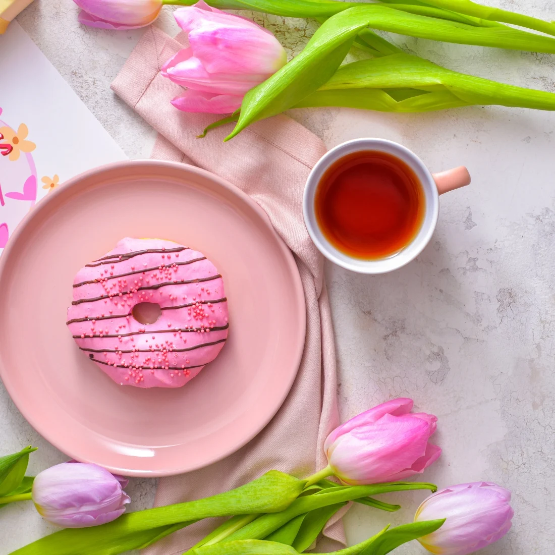 Pink frosted donut with sprinkles on a pink plate, cup of tea, and pink tulips arranged on a light surface. A pink napkin and a floral card are also visible in the scene.