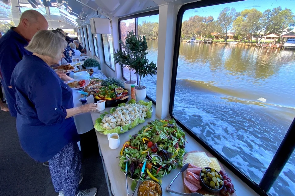 People selecting food from a buffet table on a boat. The table is filled with salads, fruits, and cheeses. Large windows offer a view of calm water and trees, creating a relaxing atmosphere.