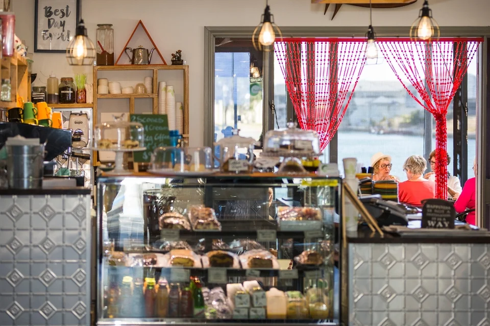 A cosy café interior with a display case filled with pastries and desserts. Hanging lights illuminate the counter. A red curtain frames the window, through which two people can be seen sitting outside. Shelves hold various items and decor.