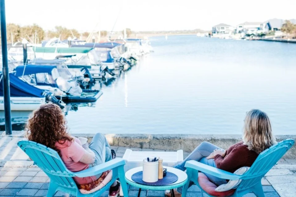 Two people sit on turquoise chairs facing a tranquil marina filled with boats. They are on a patio with a small table between them, enjoying the sunny day and peaceful water view. Houses are visible in the background along the waterfront.