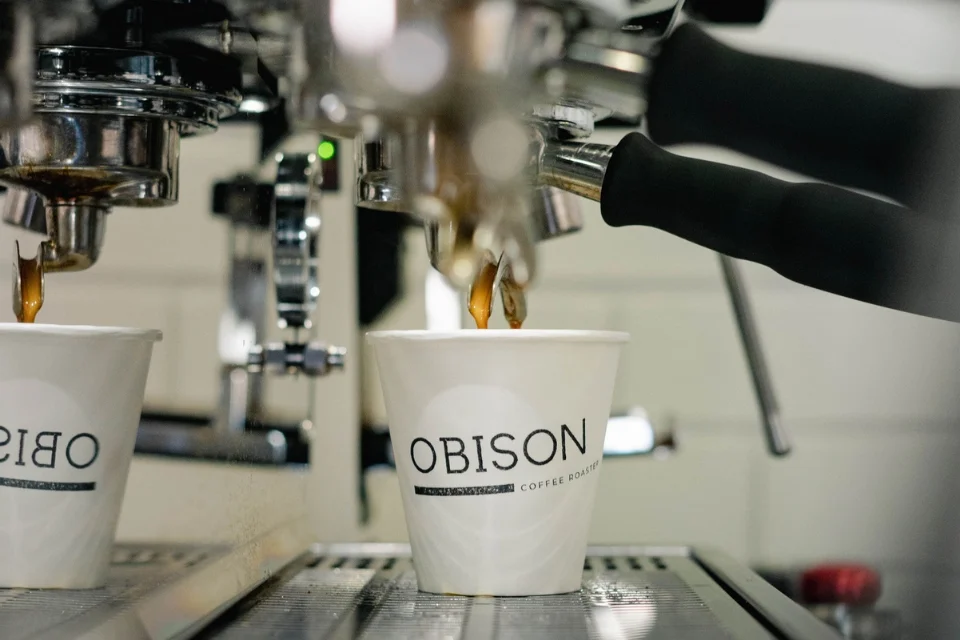 Close-up of an espresso machine brewing coffee into a white paper cup on a metallic tray. The cup features the text OBISON in bold letters. Two black handles of the espresso machine are visible.