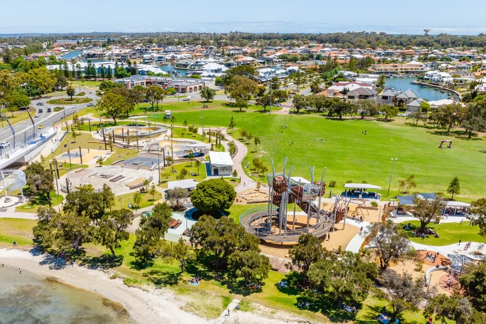 Aerial view of a coastal park with a modern playground, green open spaces, and walking paths. Surrounding the park are residential areas with houses and boats docked in canals. The shoreline is visible with a sandy beach, trees, and parked cars.