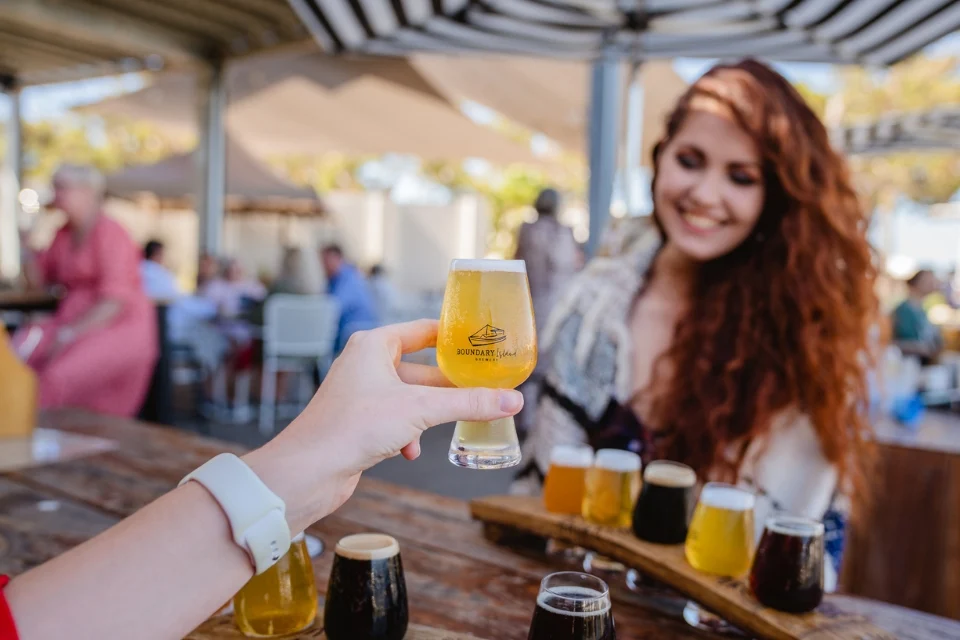 A person holds up a small glass of beer for a toast at an outdoor table. Another person with long red hair smiles, and a flight of assorted beers is on the table. The setting is a patio with umbrellas and seated patrons in the background.