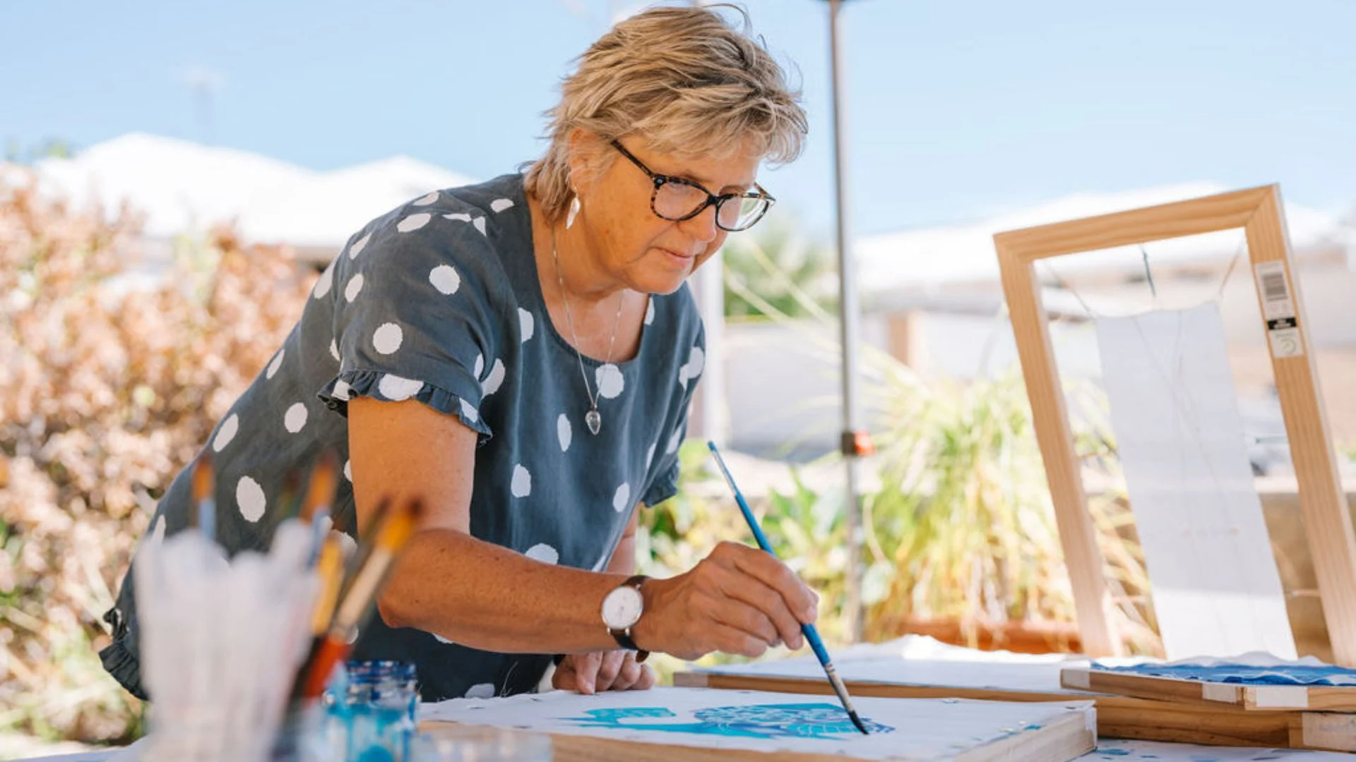 A woman wearing glasses and a polka-dot shirt paints on a canvas outdoors. She is focused on her artwork, surrounded by art supplies, with plants and bright sunlight in the background.