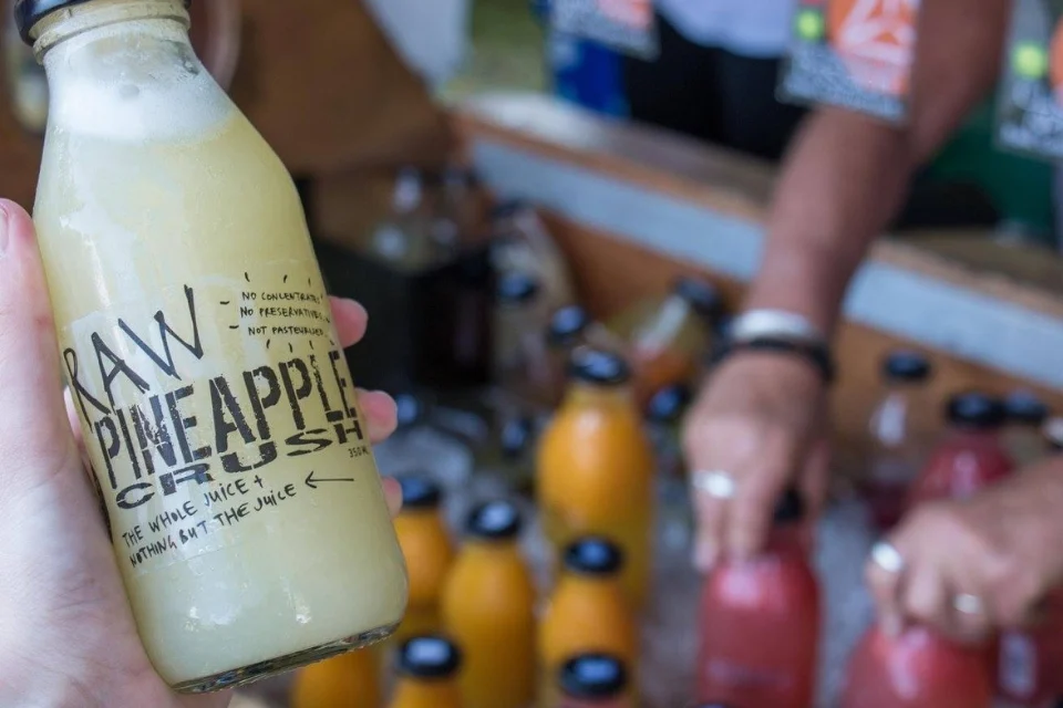 A person holds a bottle labeled Raw Pineapple Crush. In the background, a vendor serves various colorful juice bottles at a market stall. The setting appears lively and informal.