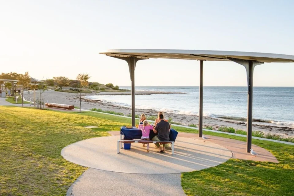 A group of people sitting at a picnic table under a modern metal canopy by the beach. They are surrounded by grass and a paved path, with the ocean and a sandy shoreline in the background under a clear sky.