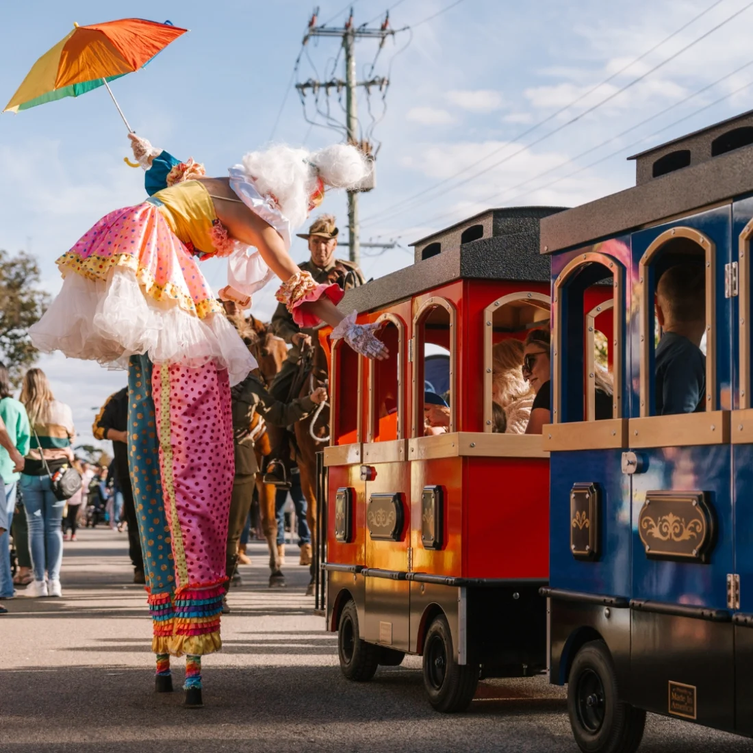 A performer on colourful stilts and holding an orange umbrella greets children riding in a small, colorful train during an outdoor festival. People and horses are visible in the background.