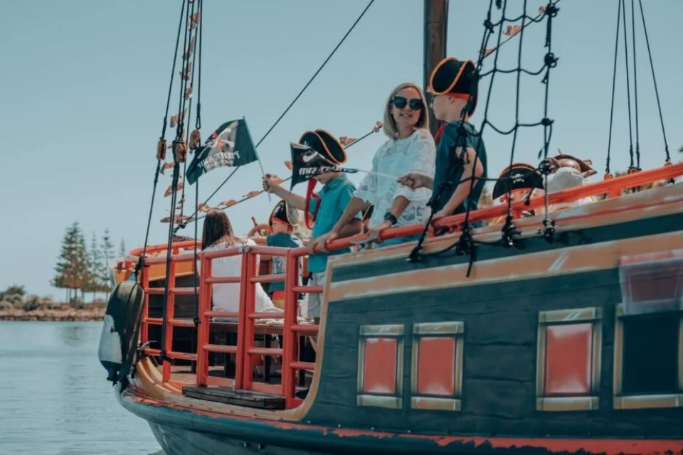 A group of children wearing pirate hats and waving a flag stand on a pirate-themed ship. An adult is with them on deck. The ship is on calm water with trees visible in the background under a clear blue sky.