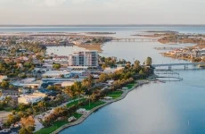 Aerial view of the coastal town of Mandurah with buildings, green spaces, and trees along a waterfront, with bridges and calm blue water extending into the distance.