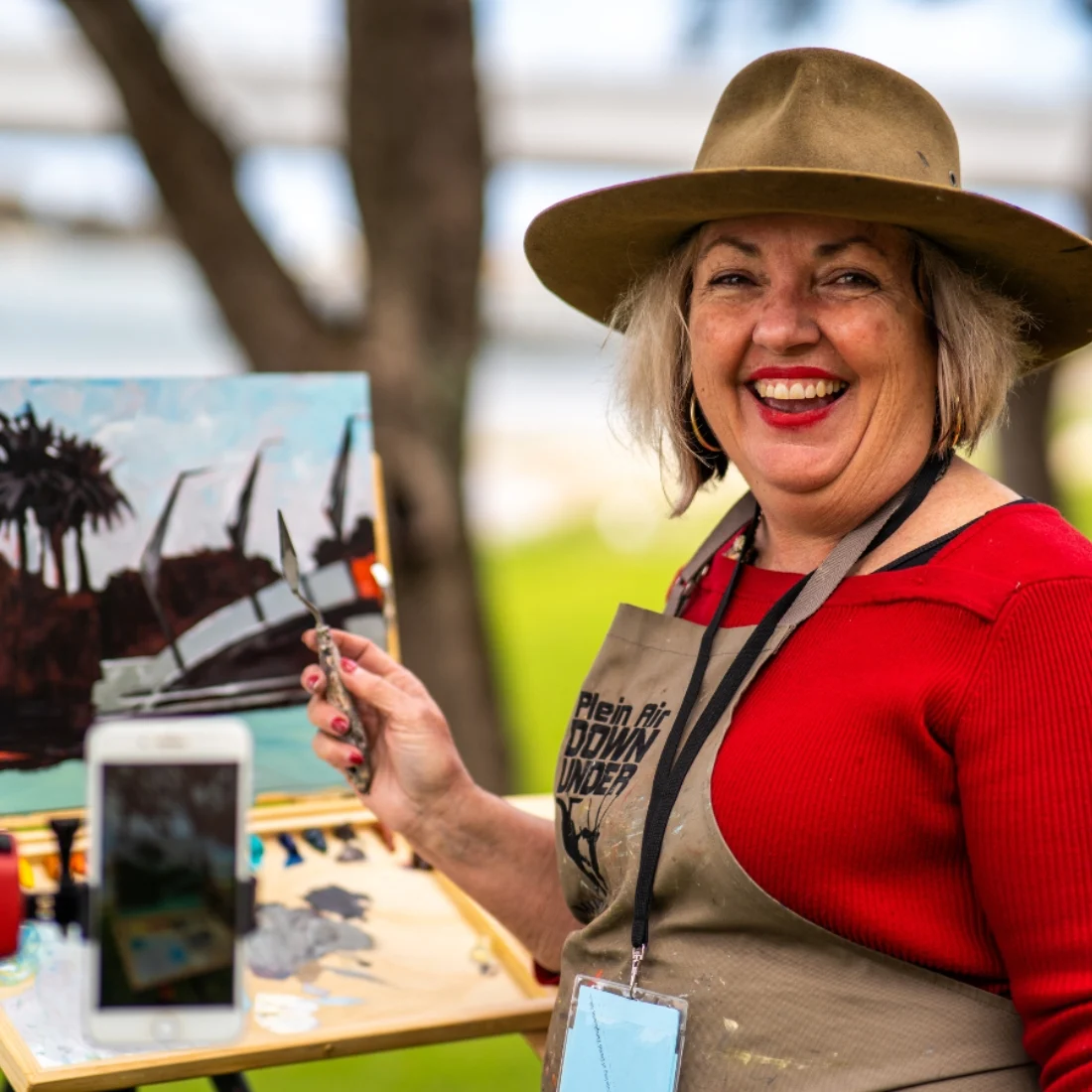 A smiling woman in a wide-brimmed hat and red top holds a paintbrush next to an easel with a partially finished landscape painting outdoors. Art supplies and a phone are on the table beside her.