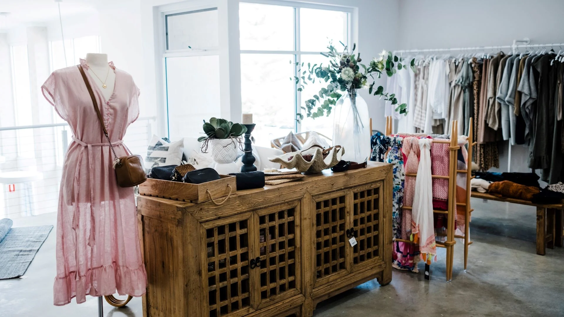 A clothing boutique interior with a pink dress displayed on a mannequin. A wooden cabinet holds bags and decor items. Racks in the background showcase various garments in neutral tones, alongside a display of colourful scarves.