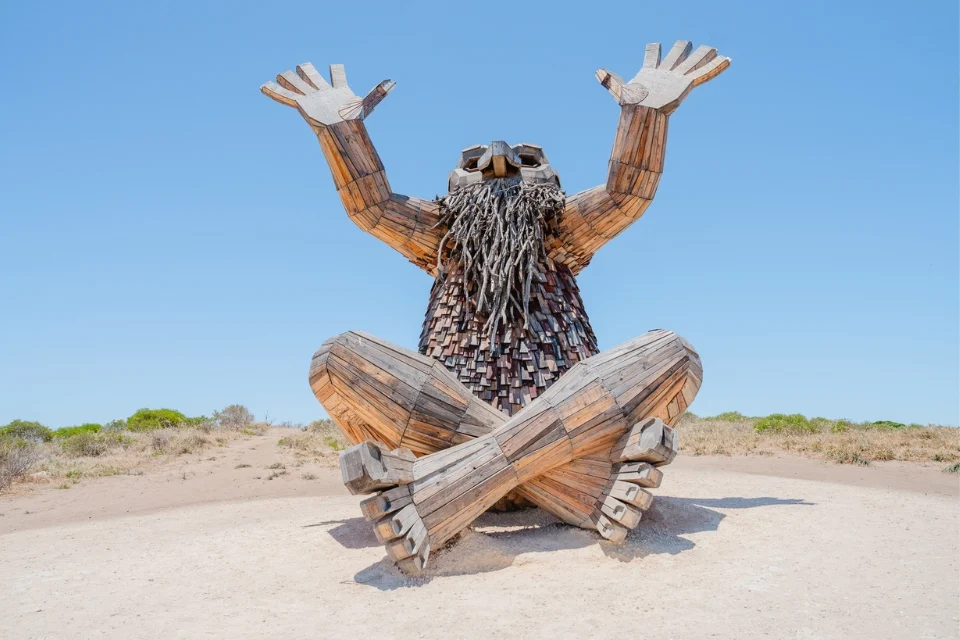 A large wooden sculpture of a bearded figure sits cross-legged with arms raised to the sky. The artwork is made from various pieces of wood and sits on a sandy terrain against a clear blue sky.