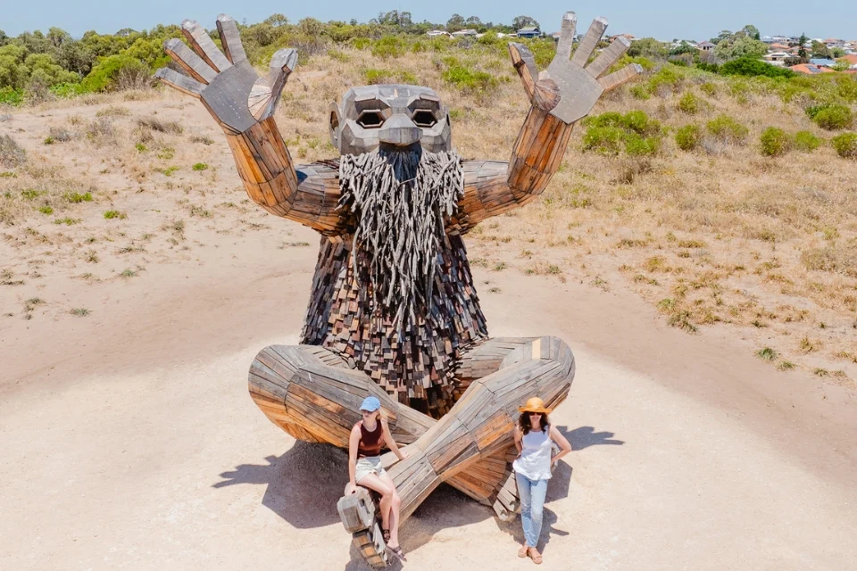 A large wooden sculpture of a seated humanoid figure with outstretched hands and a long beard occupies an outdoor setting. Two people are interacting with the sculpture, one sitting on its foot and the other standing nearby. Its a sunny day.