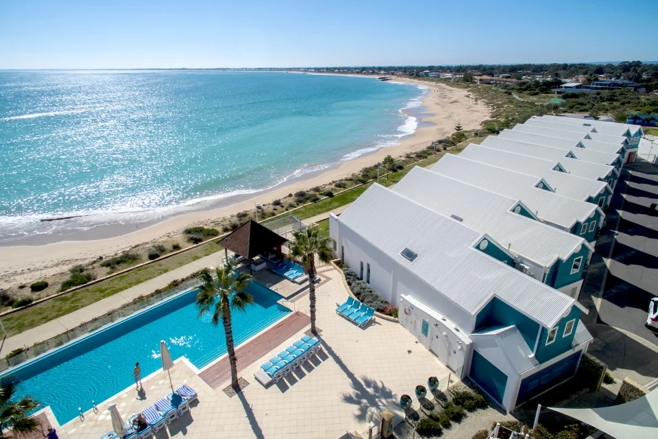 Aerial view of a coastal resort featuring a white building with light blue accents. A turquoise swimming pool is surrounded by palm trees and lounge chairs. The expansive beach and ocean extend along the shoreline under a clear blue sky.