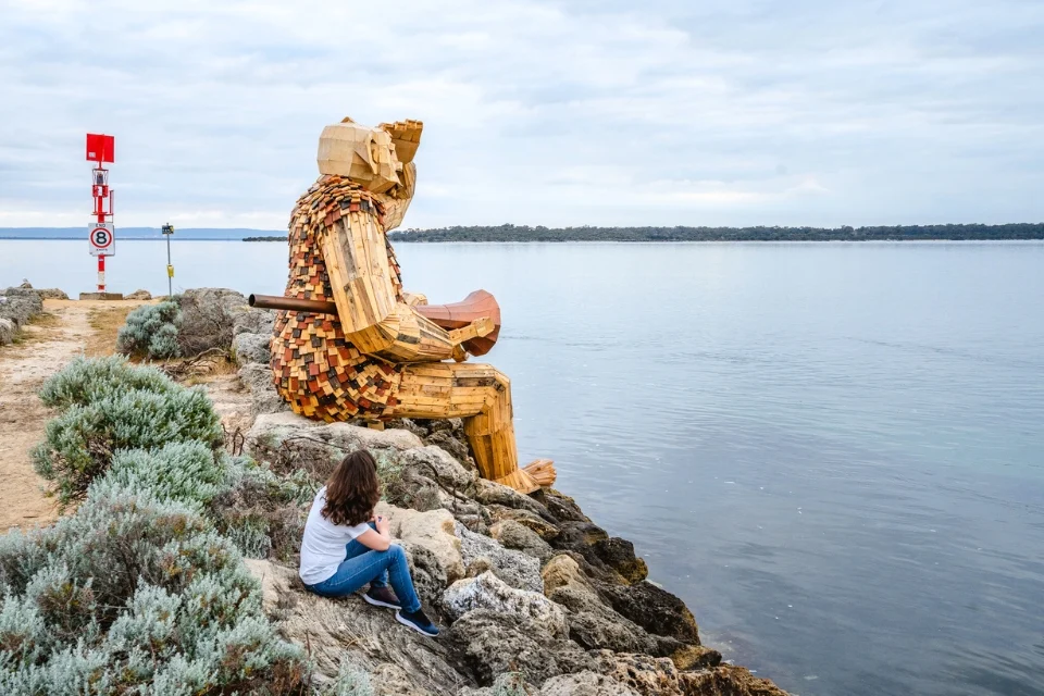 A giant wooden sculpture of a seated figure overlooks a calm body of water, resting on a rocky shoreline. A person with long hair, wearing a white shirt and jeans, sits nearby on the rocks. A cloudy sky extends over the scene.