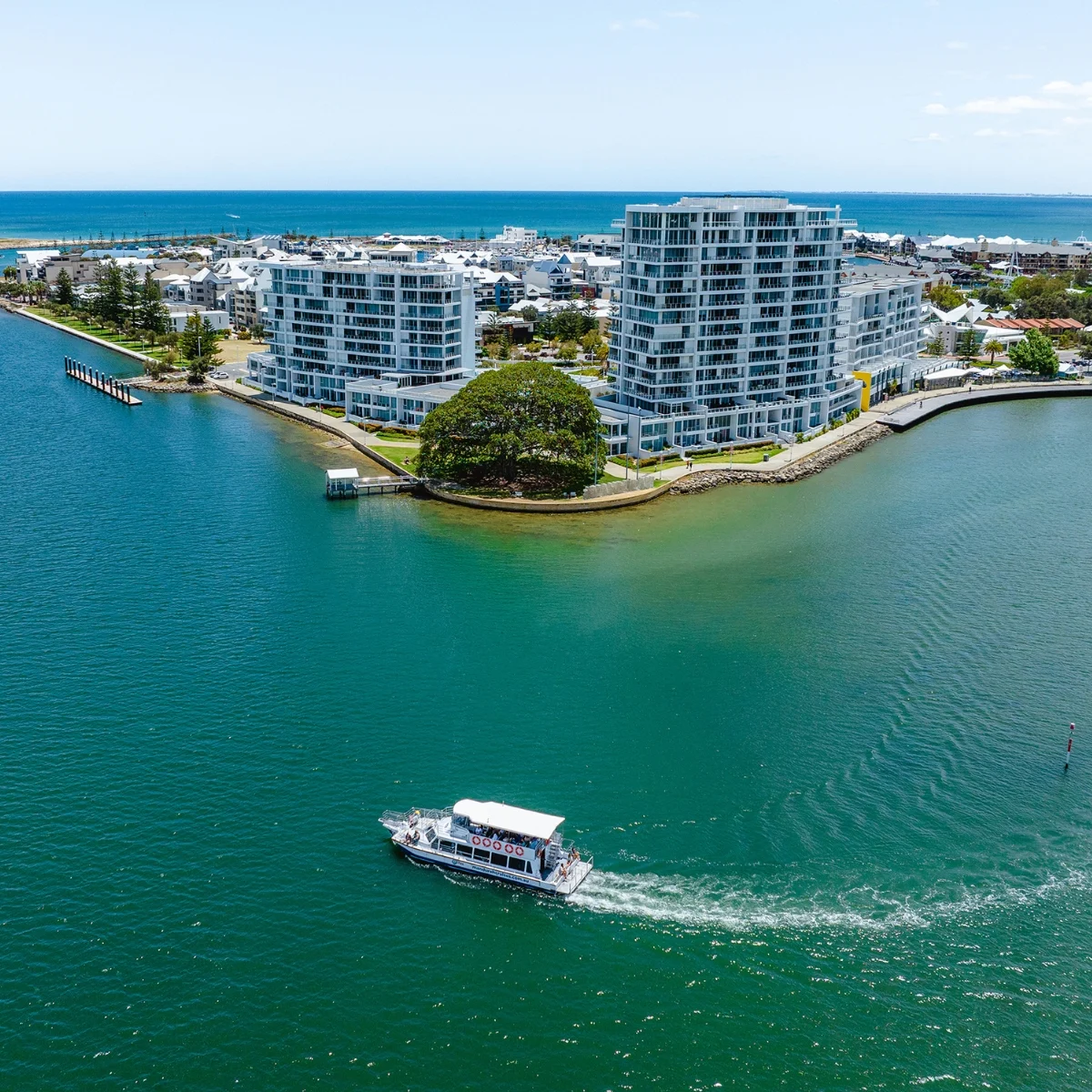 A small boat travels across blue-green water near modern waterfront apartment buildings, with a large leafy tree and a distant view of the ocean and sky in the background.