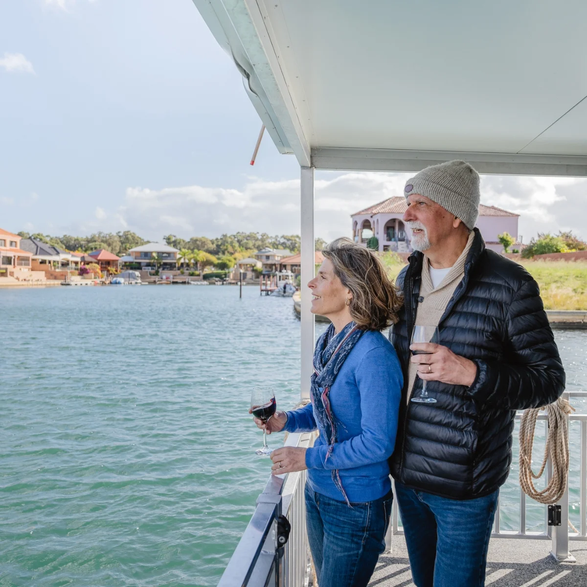 An older couple stands on a houseboat deck by the water, smiling and holding glasses of wine while looking at waterfront houses on a sunny day.