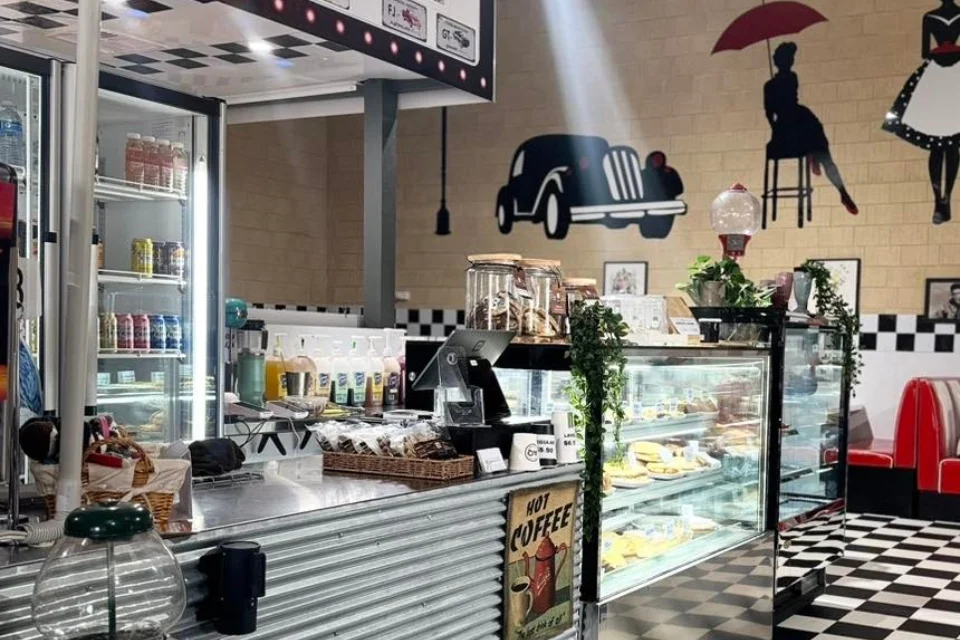 Retro-themed café with checkered floors and a vintage car mural. A display counter holds pastries and snacks, while a fridge is stocked with drinks. Red booths and a wall silhouette of a woman with an umbrella add to the nostalgic atmosphere.