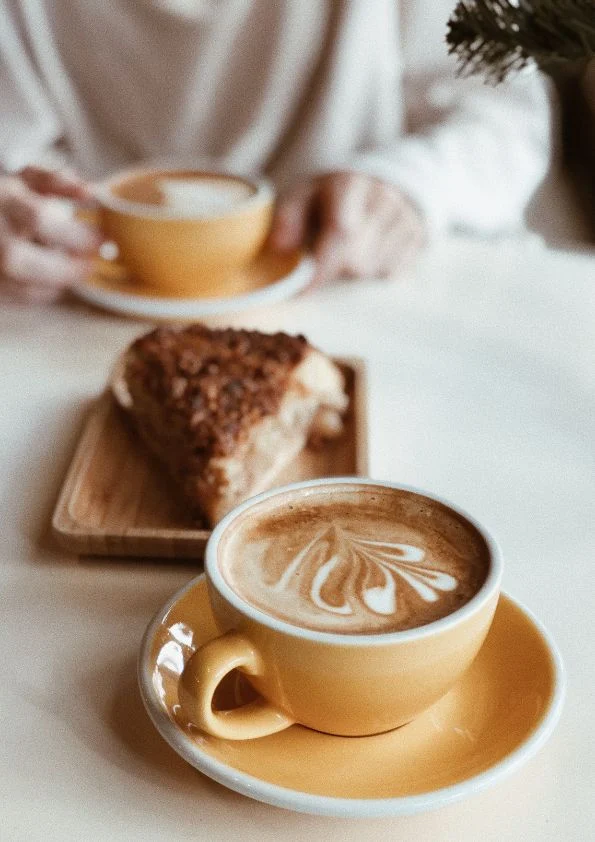A close-up of a yellow cup of coffee with latte art, next to a slice of cake on a wooden tray. In the background, a person holds another yellow coffee cup. The setting appears to be a cosy café.