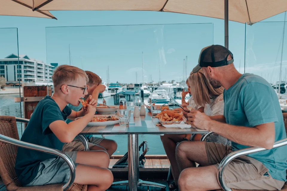 A family of four is dining outdoors at a marina restaurant. They are seated around a table enjoying burgers and fries. The background shows yachts docked in the harbor under a clear blue sky.