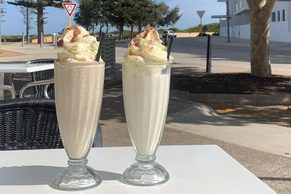 Two tall milkshakes topped with whipped cream and cocoa sit on an outdoor cafe table. Theres a street in the background with a stop sign, sidewalks, and trees under a clear sky.