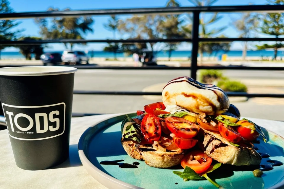 A brunch scene with a plate of toast topped with tomatoes, greens, and a poached egg, drizzled with balsamic glaze. A coffee cup labeled TODS sits beside. The background shows a road, trees, and the ocean under a clear blue sky.