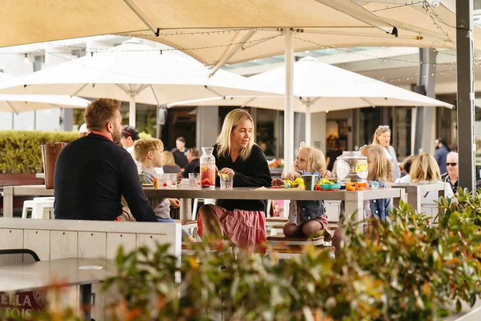 People sitting at a large outdoor table enjoying a meal under white umbrellas. A woman interacts with a child while others converse nearby. The setting is casual and lively with greenery surrounding the area.
