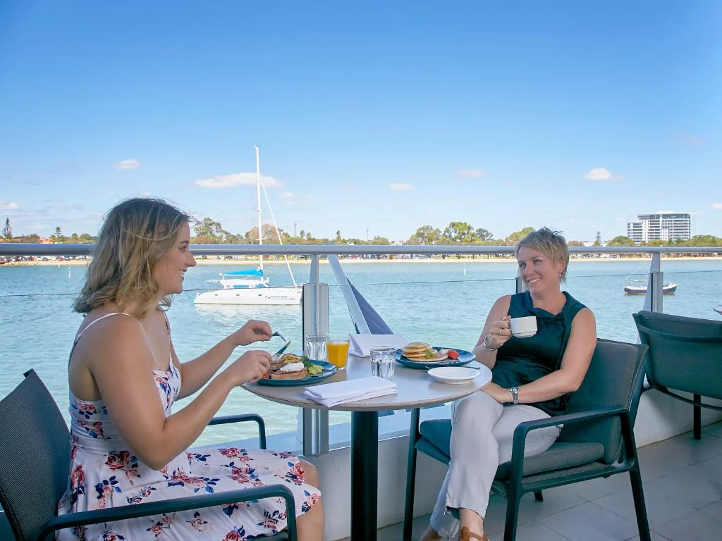 Two people enjoy a meal on an outdoor patio overlooking a serene body of water. The table is set with plates of food and a glass of orange juice. A sailboat is visible in the water, and the sky is clear and blue.