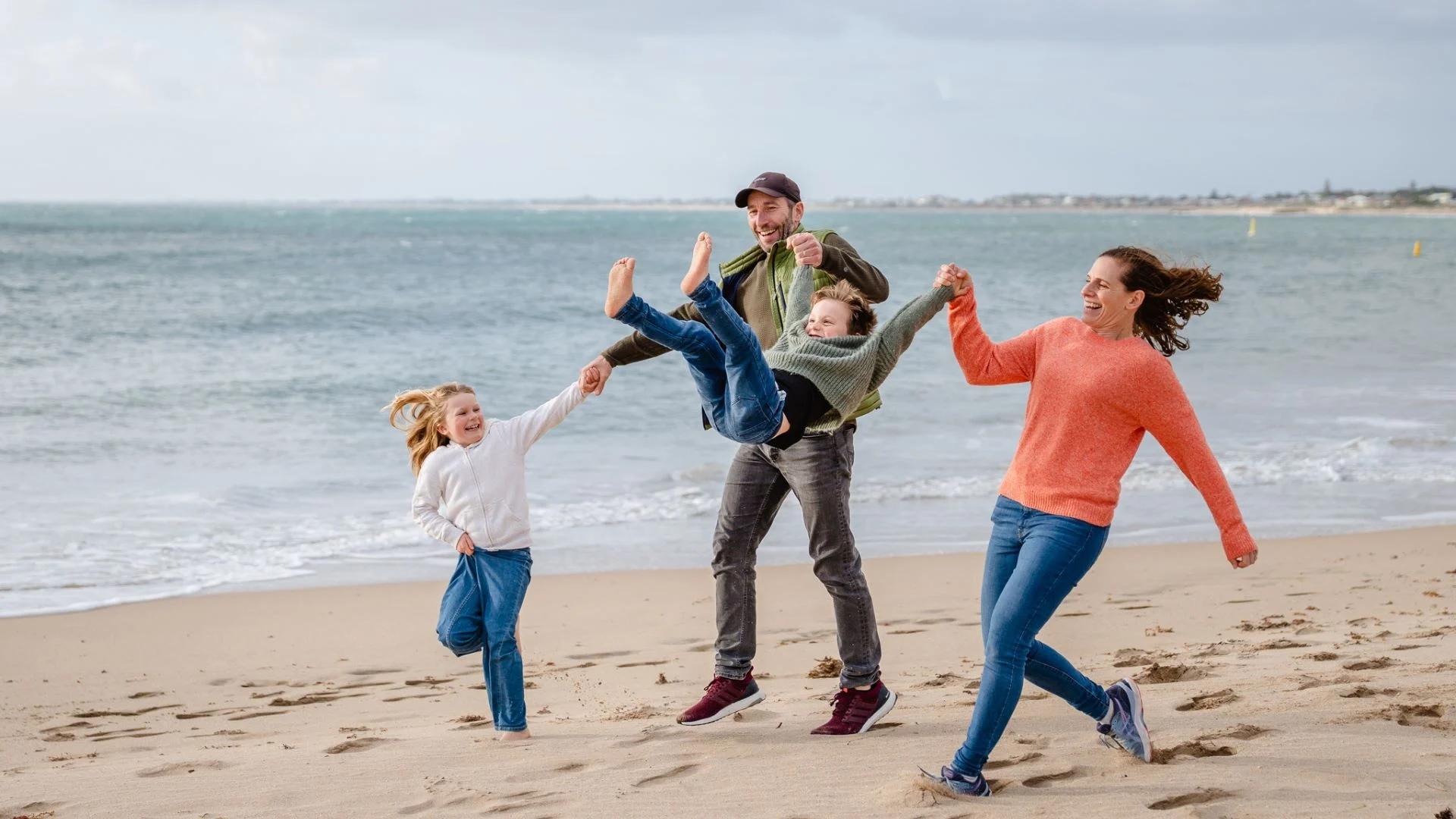 A family of four joyfully plays on a beach. Two adults hold a child who is kicking their legs in the air. Another child runs beside them. The ocean and a distant shoreline are in the background under a cloudy sky.