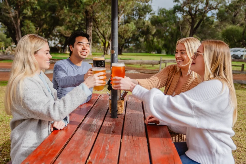 Four people sitting at a red picnic table outdoors, toasting with beer glasses. Theyre smiling and appear to be enjoying a sunny day in a park-like setting with trees in the background.