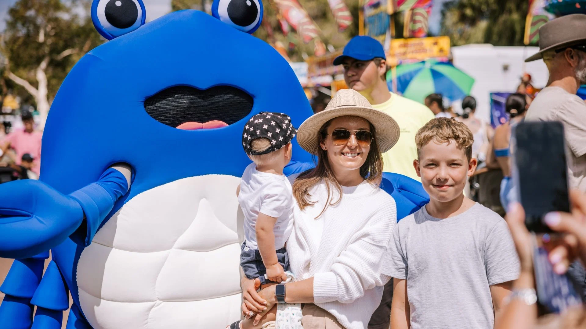 A person wearing a blue crab costume poses with a woman in sunglasses and a hat holding a toddler, and a boy in a gray shirt. They are outdoors at a festive event, with colorful flags and a crowd in the background.