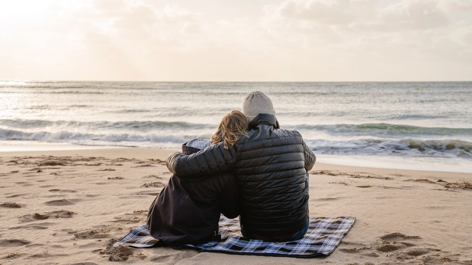 A couple sits on a plaid blanket at the beach, embracing each other while facing the ocean. They are wearing jackets and a beanie, suggesting a cool day. The sky is cloudy but bright, with the sun setting near the horizon over the calm sea.